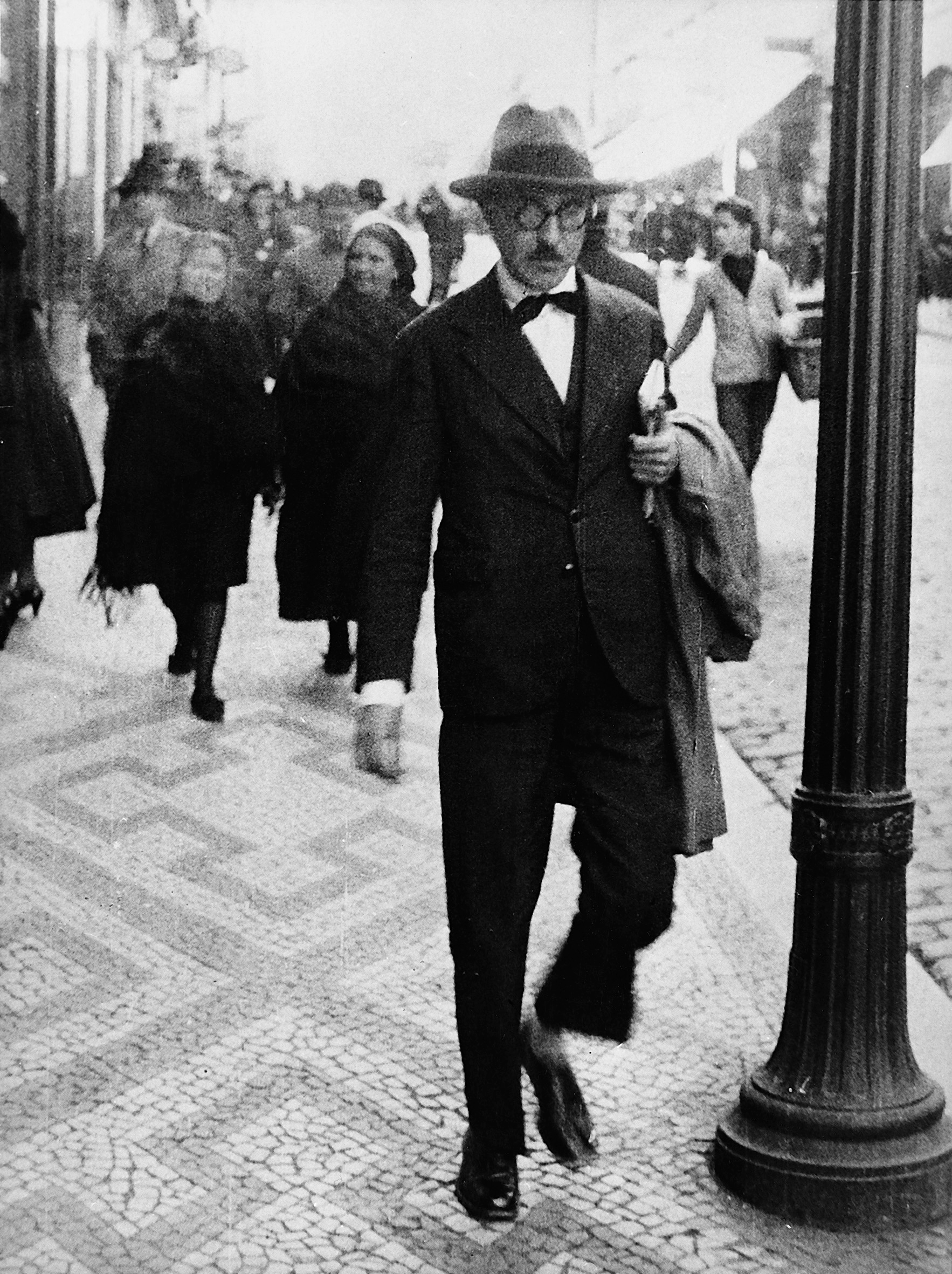 black-and-white photo of portuguese author Fernando Pessoa walking through a street in Lisbon, 1920s or 1930s, with other passersby in the background