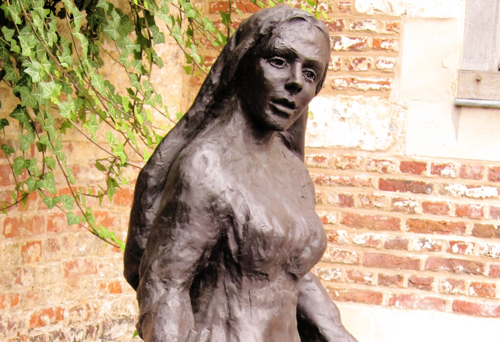 bronze (or similar) statue of a beguine woman (a semi-monastic community in the 13th-16th century) in the courtyard (with red brick walls and greenery in the background) of a beguinage in Tongern, Belgium